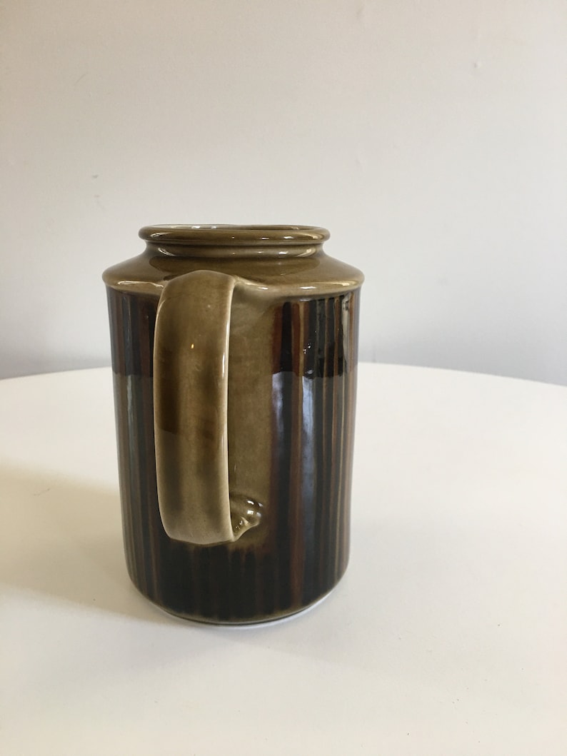 Puede incluir: Una jarra de cer&aacute;mica marr&oacute;n y beige con un dise&ntilde;o a rayas. La jarra tiene un asa y una boca ancha.