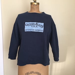 May include: A navy blue sweatshirt with a light blue embroidered patch that says "Guess Jeans U.S.A. Satisfaction Guaranteed".