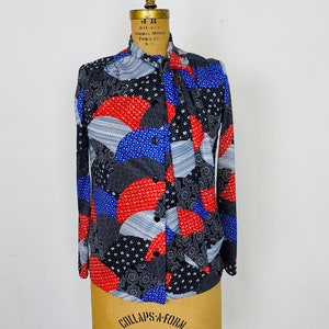 May include: A long-sleeved button-up shirt with a geometric pattern in shades of blue, red, white, and black. The shirt has a collar and a tie at the neck.