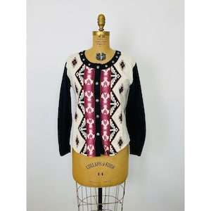 May include: A long-sleeved cardigan with a cream-colored front panel featuring a geometric pattern in burgundy, black, and white. The sleeves are solid black, and the cardigan has silver button closures.