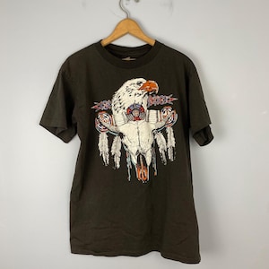 Puede incluir: Una camiseta marrón oscuro con un estampado gráfico de un águila blanca con un diseño rojo, blanco y azul. El águila está posada sobre un cráneo con plumas y cuentas.