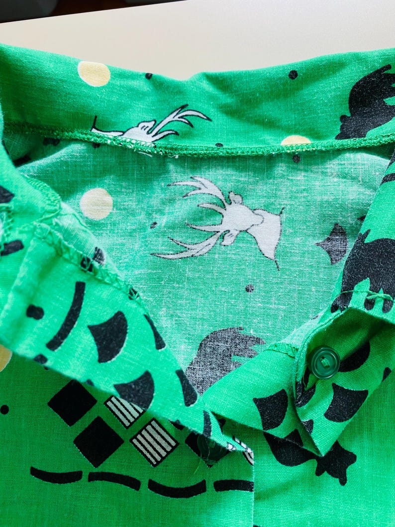 Puede incluir: Primer plano de una camisa verde con cuello con un patr&oacute;n repetido de ciervos blancos, osos negros, c&iacute;rculos amarillos y formas geom&eacute;tricas. La camisa tiene cierre de bot&oacute;n y est&aacute; hecha de tela ligera.