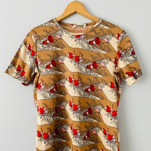 May include: Short-sleeved t-shirt with a repeating pattern of figures in red hats and umbrellas against a tan and cream landscape. The shirt is hanging on a wooden hanger against a white background.