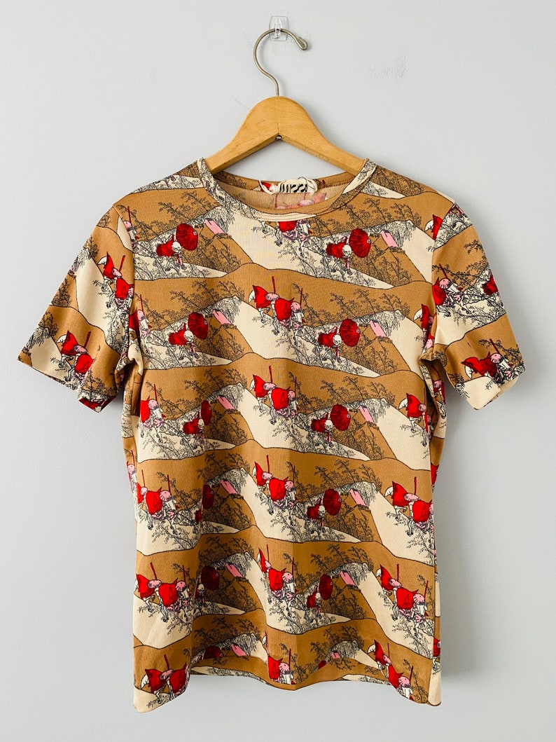 May include: Short-sleeved t-shirt with a repeating pattern of figures in red hats and umbrellas against a tan and cream landscape. The shirt is hanging on a wooden hanger against a white background.