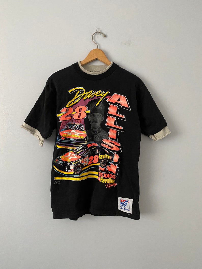 May include: Black t-shirt featuring a colourful graphic of race car driver Davey Allison, his car, and the text "Davey Allison 28 Texaco Havoline Racing".