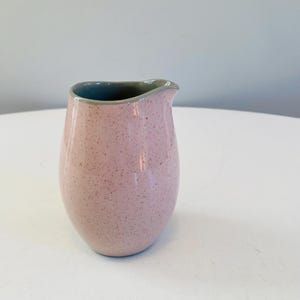 Op de afbeelding: Een kleine roze keramische roomkan met een grijze rand. De roomkan heeft een gespikkelde textuur.