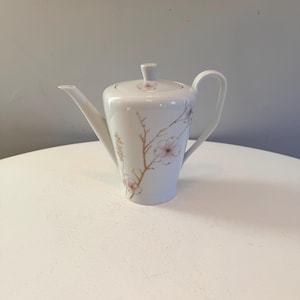May include: White porcelain teapot with a lid and a curved handle. The teapot is decorated with delicate floral branches in shades of gold and pink. The spout is long and slender, and the overall design is elegant and refined.