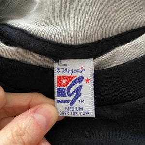 May include: A black t-shirt with a white and grey ribbed collar. The tag on the shirt reads "The Game" with a stylized "G" logo and the words "Medium" and "Over for Care".