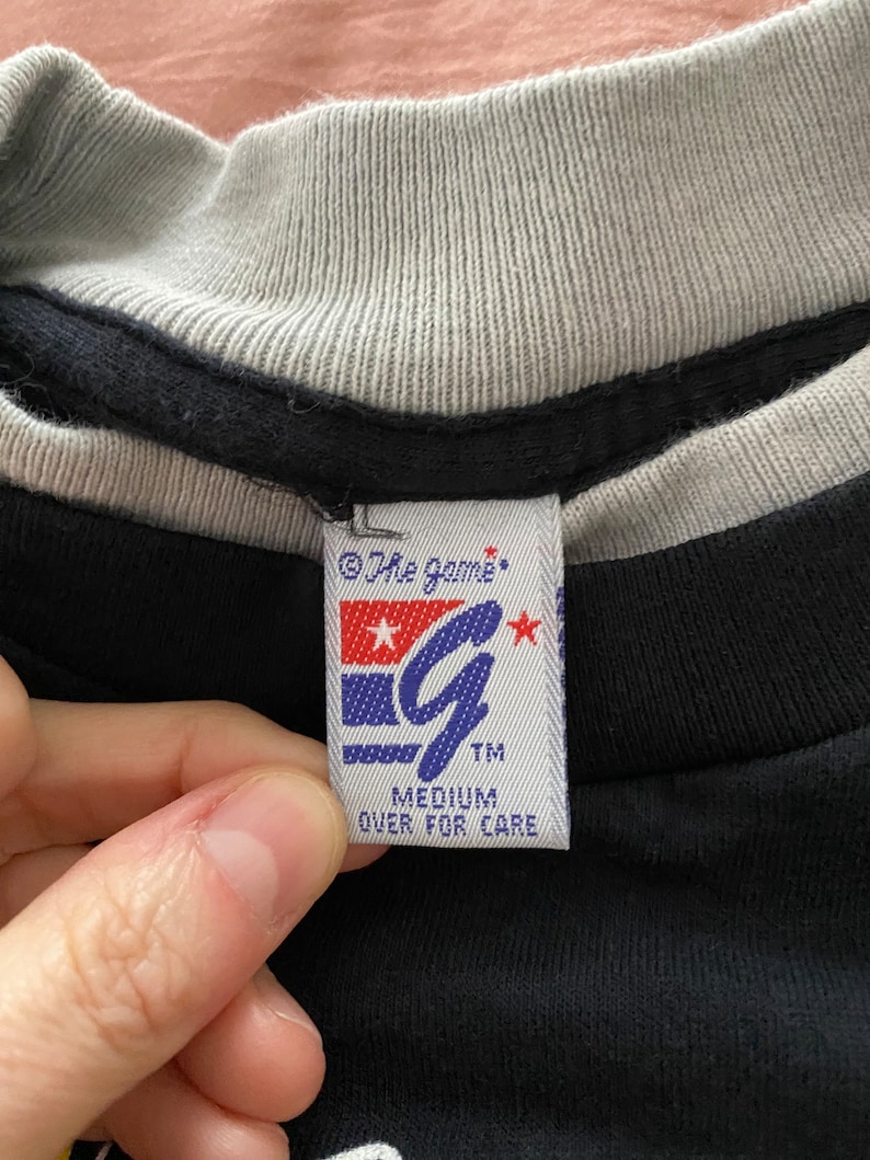 May include: A black t-shirt with a white and grey ribbed collar. The tag on the shirt reads "The Game" with a stylized "G" logo and the words "Medium" and "Over for Care".