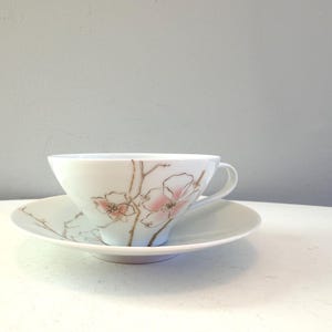 Op de afbeelding: Witte theekop en schotel set met een bloemenontwerp. De kop en schotel zijn versierd met roze bloemen en bruine stelen. De theekop heeft een gebogen oor en staat op een bijpassende schotel. Een delicate en elegante theeset.