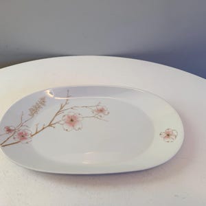 May include: Oval white serving platter with a floral design. The platter features a delicate branch with pink blossoms and a smaller blossom on the side. The design is in shades of pink and brown.