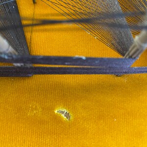 May include: A close-up of a wooden loom with black string stretched across it. The loom is sitting on a yellow fabric surface with a small hole in it.