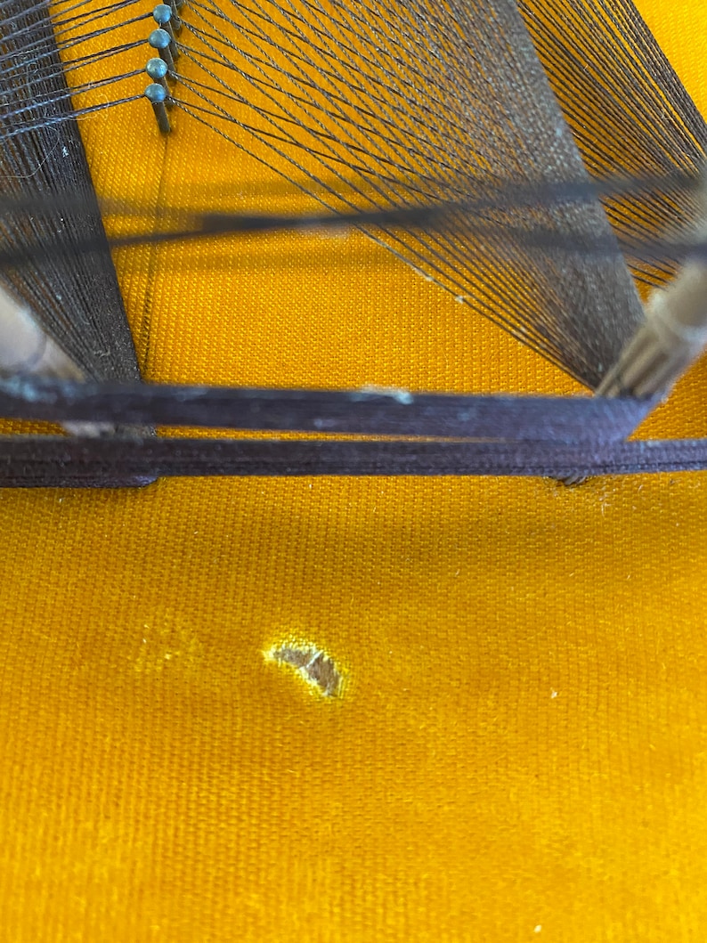 May include: A close-up of a wooden loom with black string stretched across it. The loom is sitting on a yellow fabric surface with a small hole in it.