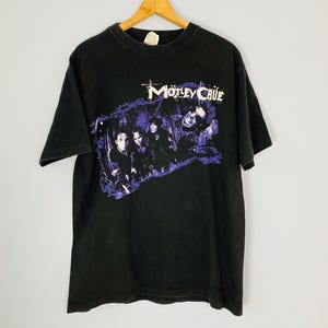 Pode incluir: Camiseta preta com a banda Mötley Crüe. A camisa exibe o nome da banda em branco e um gráfico roxo dos membros da banda. A camiseta está pendurada em um cabide de madeira.