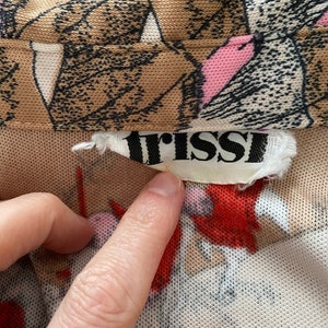 May include: Close-up of a beige mesh fabric with a floral pattern in brown, pink, and red. The label reads "trissi" in black letters. A finger points to the label.
