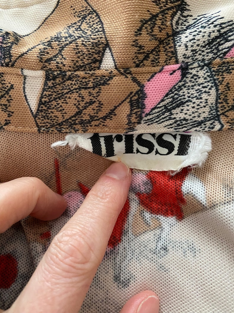 May include: Close-up of a beige mesh fabric with a floral pattern in brown, pink, and red. The label reads "trissi" in black letters. A finger points to the label.