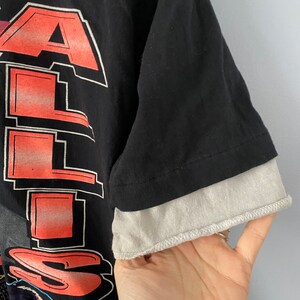 May include: Black t-shirt with a red and white graphic print of the word "ALL" and a cartoon image of a person riding a motorcycle. The shirt has a grey underlay on the sleeves.