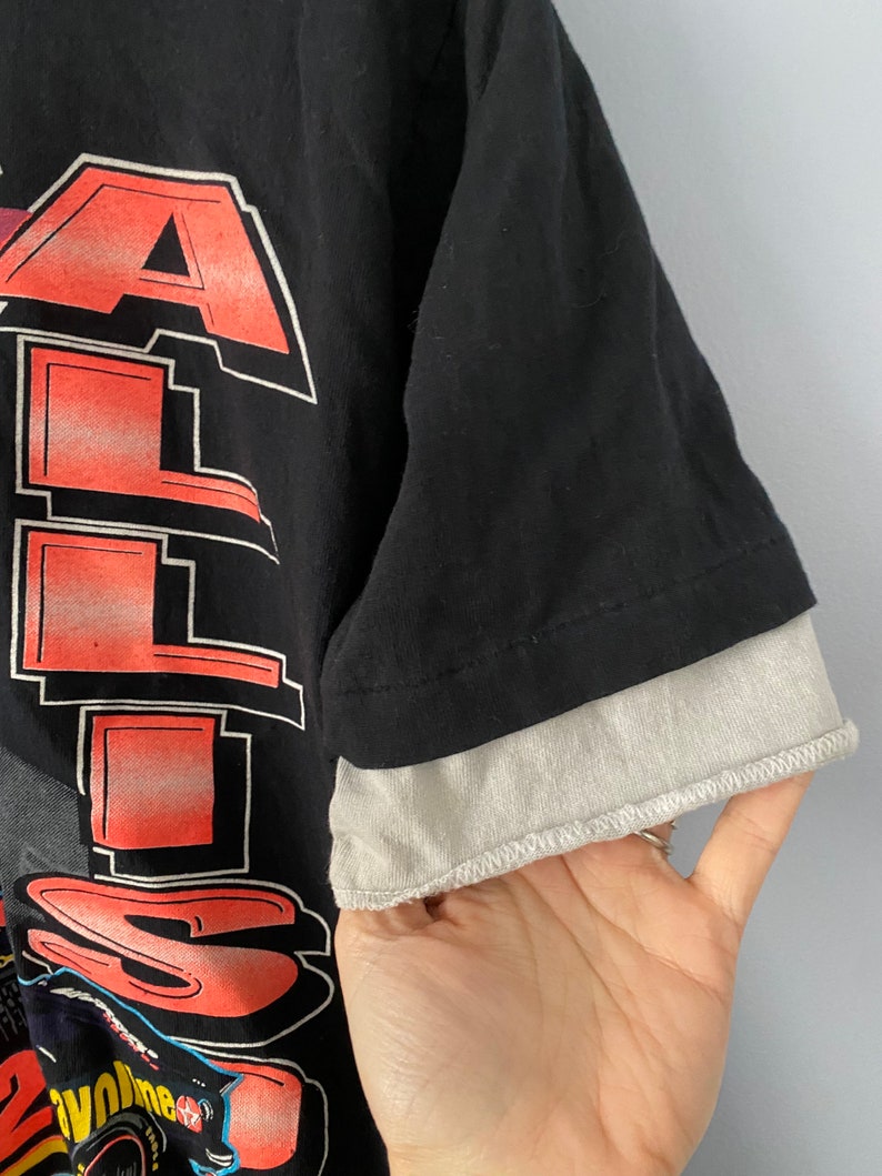 May include: Black t-shirt with a red and white graphic print of the word "ALL" and a cartoon image of a person riding a motorcycle. The shirt has a grey underlay on the sleeves.