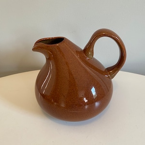 May include: A brown ceramic pitcher with a rounded body and a curved handle. The pitcher has a speckled glaze.