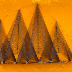 May include: A wall hanging made of string stretched between nails, forming a geometric pattern of triangles. The string is a dark brown color, and the nails are silver. The background is a bright yellow.