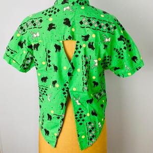 Puede incluir: Camisa verde brillante de manga corta con un dise&ntilde;o &uacute;nico de espalda abierta. La camisa presenta un patr&oacute;n repetido de siluetas de animales negros, p&aacute;jaros blancos y puntos amarillos. La tela tiene una apariencia ligeramente texturizada.