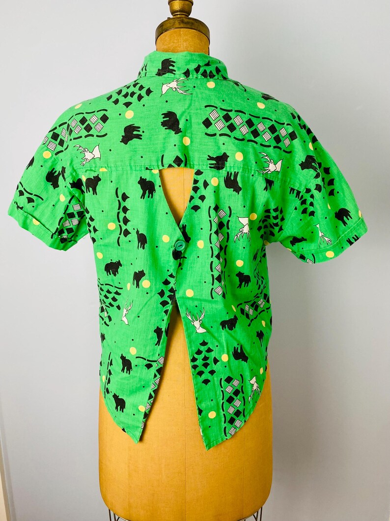 Puede incluir: Camisa verde brillante de manga corta con un dise&ntilde;o &uacute;nico de espalda abierta. La camisa presenta un patr&oacute;n repetido de siluetas de animales negros, p&aacute;jaros blancos y puntos amarillos. La tela tiene una apariencia ligeramente texturizada.