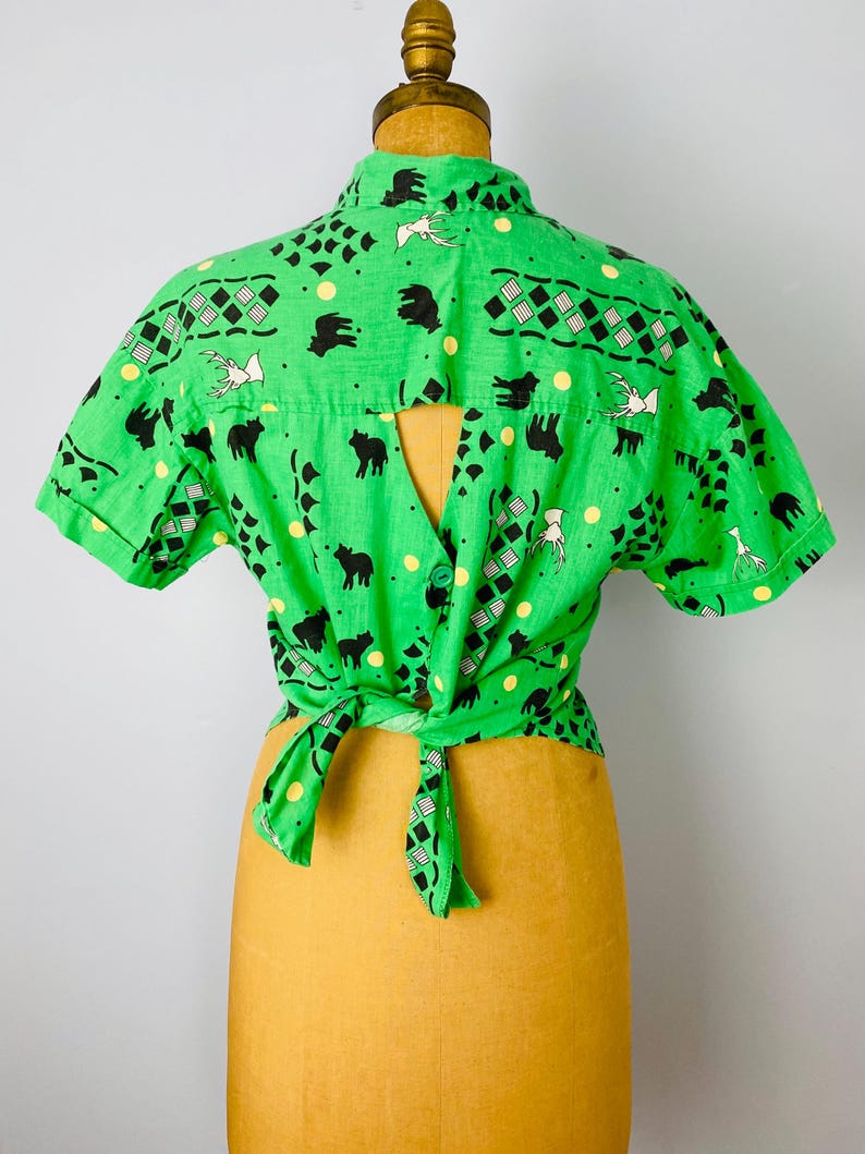 Puede incluir: Una camisa verde brillante con mangas cortas, con un patr&oacute;n repetido de siluetas de animales negros, p&aacute;jaros blancos y formas geom&eacute;tricas. La camisa est&aacute; atada a la cintura y se muestra en un maniqu&iacute;.