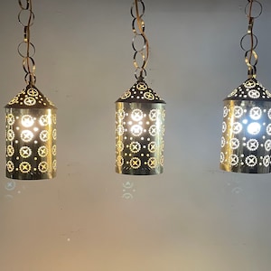 Może przedstawiać: Trzy mosiężne lampy wiszące z misternymi wzorami wyciętymi. Lampy wiszą na łańcuchach i mają ciepłe, złote światło.