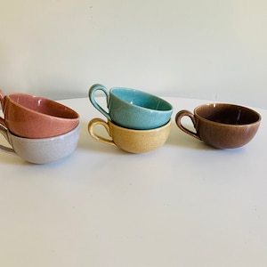 May include: Four ceramic teacups in different colors: light gray, pink, light blue, and brown. The cups are stacked in pairs, with the gray and pink cups on the left, the blue and yellow cups in the middle, and the brown cup on the right.