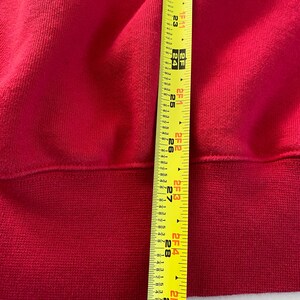 May include: A close-up of a red sweatshirt with a ribbed hem. A yellow measuring tape is visible along the bottom edge of the sweatshirt, showing a measurement of 29 inches.