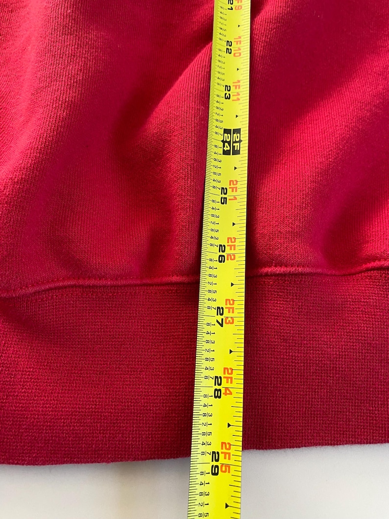 May include: A close-up of a red sweatshirt with a ribbed hem. A yellow measuring tape is visible along the bottom edge of the sweatshirt, showing a measurement of 29 inches.