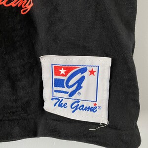 May include: Black t-shirt with a white patch featuring a blue "G" with stars and the text "The Game" in blue.