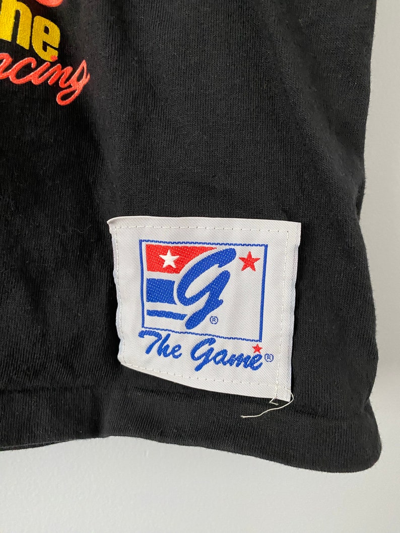 May include: Black t-shirt with a white patch featuring a blue "G" with stars and the text "The Game" in blue.