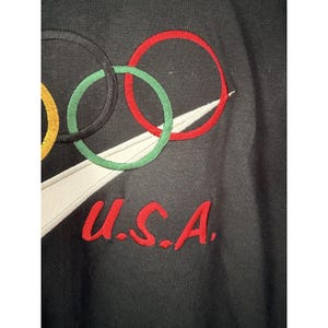 Może przedstawiać: Czarna bluza z haftowanymi pierścieniami olimpijskimi w kolorze ż&oacute;łtym, czarnym, zielonym i czerwonym. Widoczny jest biały wz&oacute;r swoosh, a także czerwone, haftowane litery "U.S.A."