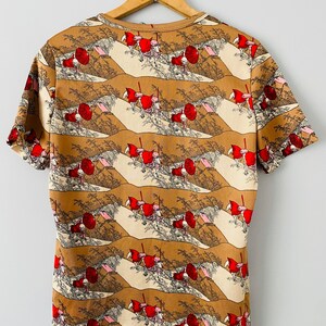 May include: A short-sleeved t-shirt with a repeating pattern of a landscape scene featuring figures in red clothing. The shirt's design includes tan, brown, and cream colors, with a round neckline. The shirt is hanging on a wooden hanger.
