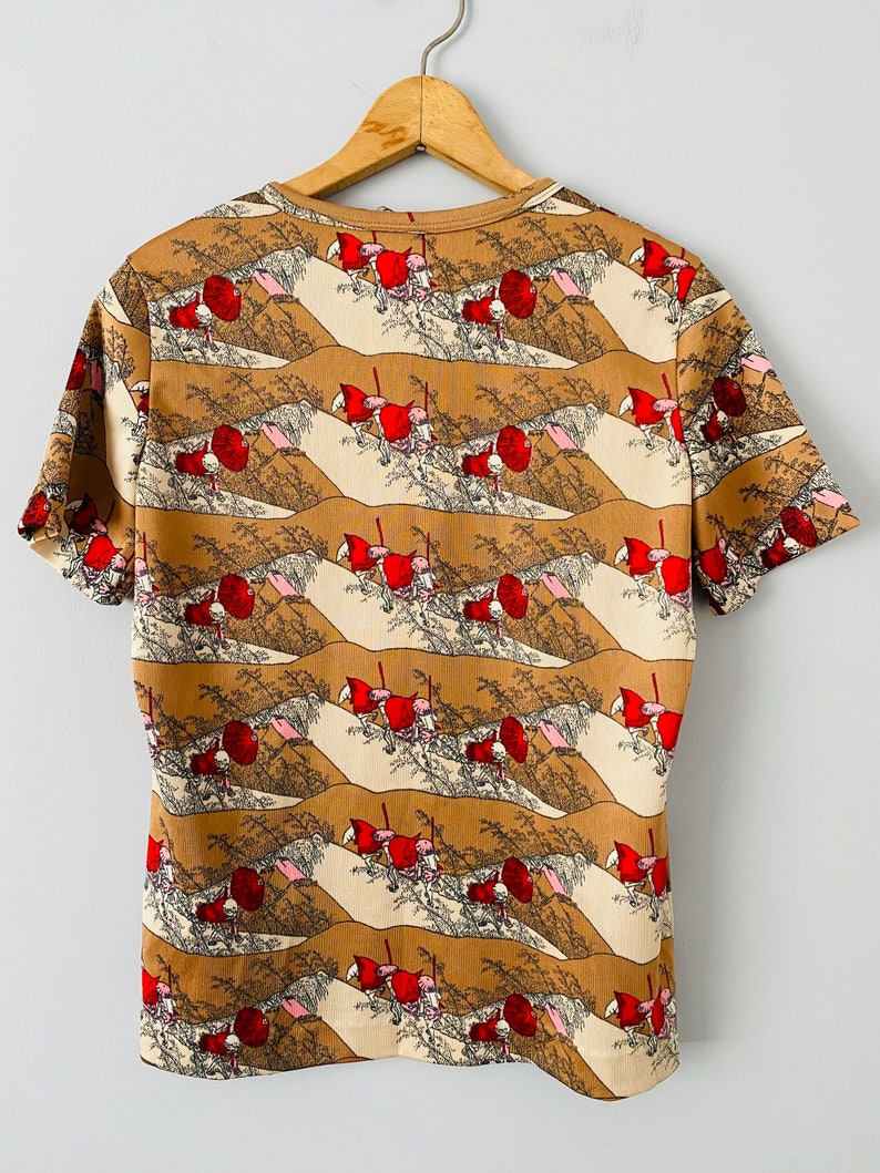 May include: A short-sleeved t-shirt with a repeating pattern of a landscape scene featuring figures in red clothing. The shirt's design includes tan, brown, and cream colors, with a round neckline. The shirt is hanging on a wooden hanger.