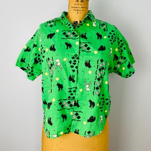 Puede incluir: Camisa verde brillante de manga corta con botones. La camisa presenta un estampado repetido de osos negros, ciervos blancos, puntos amarillos y formas geom&eacute;tricas. La camisa se exhibe en un maniqu&iacute; vintage.