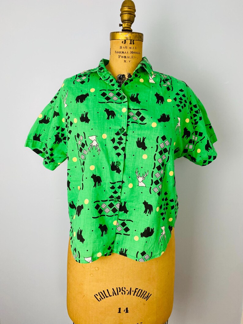 Puede incluir: Camisa verde brillante de manga corta con botones. La camisa presenta un estampado repetido de osos negros, ciervos blancos, puntos amarillos y formas geom&eacute;tricas. La camisa se exhibe en un maniqu&iacute; vintage.