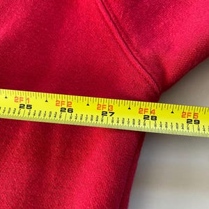 May include: Close-up of a red fabric with a yellow measuring tape. The tape reads "2F 1 25" to "2F 5 29".