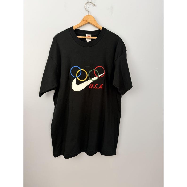 Może przedstawiać: Czarny t-shirt z logo Nike i pierścieniami olimpijskimi w kolorze niebieskim, ż&oacute;łtym, czarnym i czerwonym. Znak Nike jest biały, a napis "U.S.A." jest czerwony. Koszulka wisi na drewnianym wieszaku.