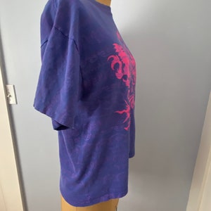 May include: A purple tie-dye t-shirt with a pink graphic of a dragon. The dragon is surrounded by a swirling pattern.