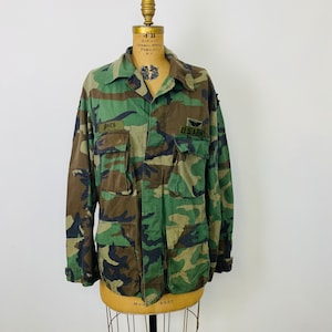 May include: A green, brown, and black camouflage jacket with a US Army patch and the word "Jones" on the left chest. The jacket is on a mannequin.