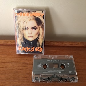 Puede incluir: Una cinta de casete del álbum "Rockbird" de Debbie Harry con los títulos de las canciones "I Want You", "French Kissin", "Buckle Up" y "In Love With Love" impresos en la caja del casete. La cinta de casete está en una caja de plástico transparente con una etiqueta negra. La etiqueta tiene el logotipo de Geffen Records y el título del álbum "Rockbird".