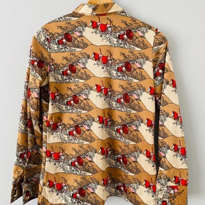 May include: A long-sleeved button-down shirt with a repeating pattern of a landscape scene in shades of brown, beige, red, and black. The shirt is hanging on a wooden hanger against a white background. The cuffs are buttoned.