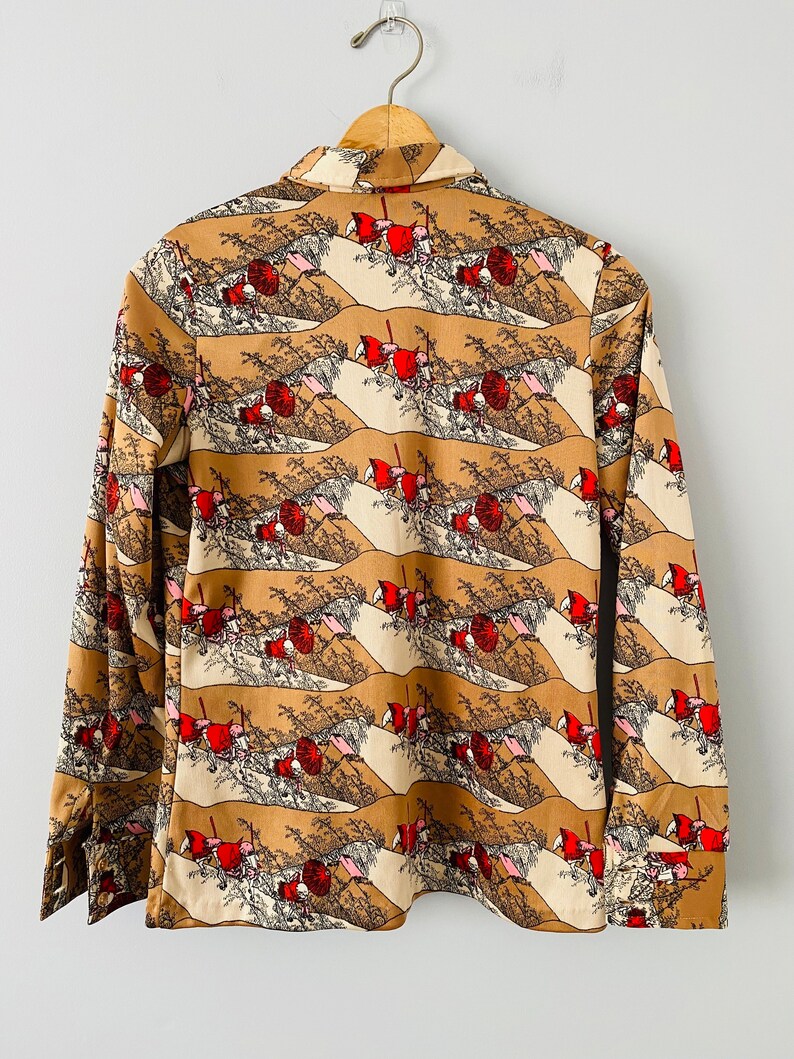 May include: A long-sleeved button-down shirt with a repeating pattern of a landscape scene in shades of brown, beige, red, and black. The shirt is hanging on a wooden hanger against a white background. The cuffs are buttoned.