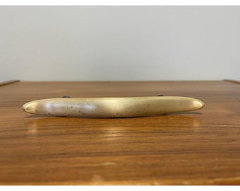 May include: A light-colored, elongated cabinet pull with a slightly curved shape. The pull has a matte finish and is mounted on a wooden surface. The hardware is designed for use on drawers or cabinet doors.