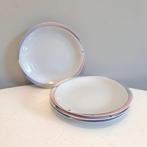 Puede incluir: Platos de cerámica blancos con un diseño de borde rosa pastel y azul. Los platos están apilados y dispuestos sobre una superficie blanca. Los platos tienen un diseño simple y elegante, adecuado para el uso diario o ocasiones especiales.