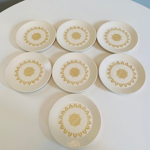 Op de afbeelding: Set van zeven witte keramische borden met een gouden bloemenontwerp. De borden zijn rond en hebben een licht verhoogde rand.