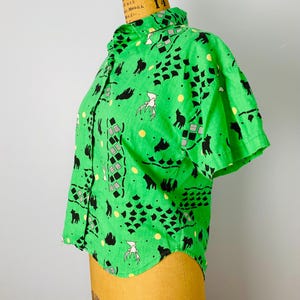 Puede incluir: Una camisa verde de manga corta con botones. La camisa presenta un patr&oacute;n repetido de siluetas de animales negros, formas geom&eacute;tricas y figuras blancas. La camisa se exhibe en un maniqu&iacute;.