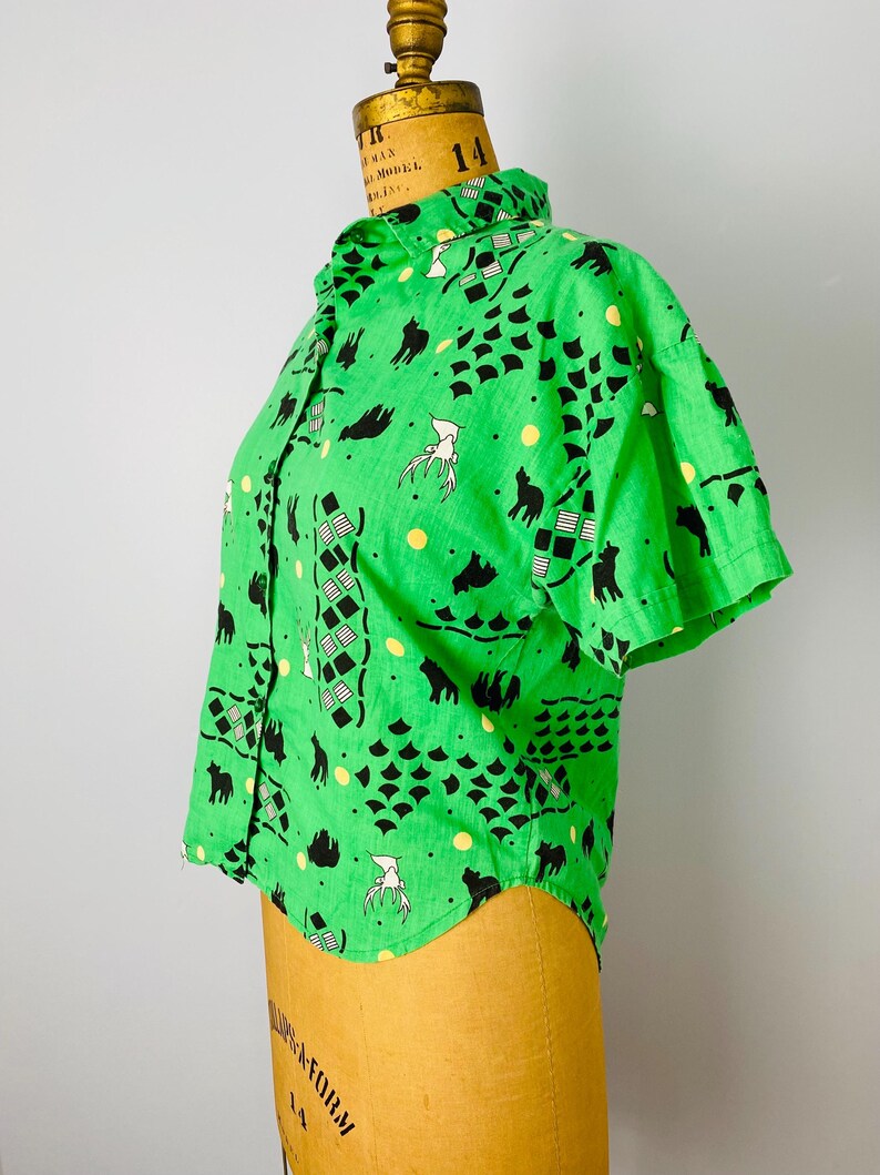 Puede incluir: Una camisa verde de manga corta con botones. La camisa presenta un patr&oacute;n repetido de siluetas de animales negros, formas geom&eacute;tricas y figuras blancas. La camisa se exhibe en un maniqu&iacute;.